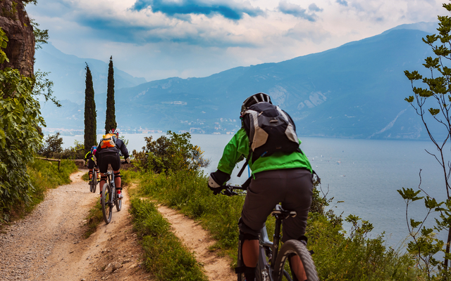 Radfahrer auf einem Pfad mit Blick auf einen See und Berge im Hintergrund.