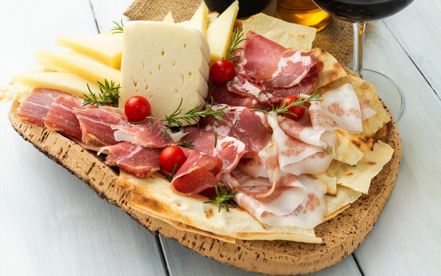 Holzplatte mit Schinken, Käse und Brot, dekoriert mit Kräutern und Tomaten.