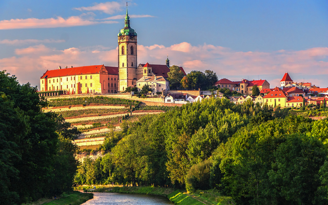 Stadtpanorama mit Schloss und Fluss im Vordergrund bei Sonnenuntergang.