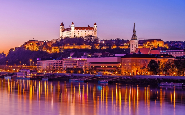 Stadtansicht von Bratislava bei Dämmerung mit Burg und Flussreflexionen.