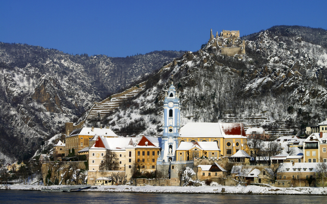 Verschneites Dorf an einem Fluss mit blauer Kirche und Burgruine im Hintergrund.