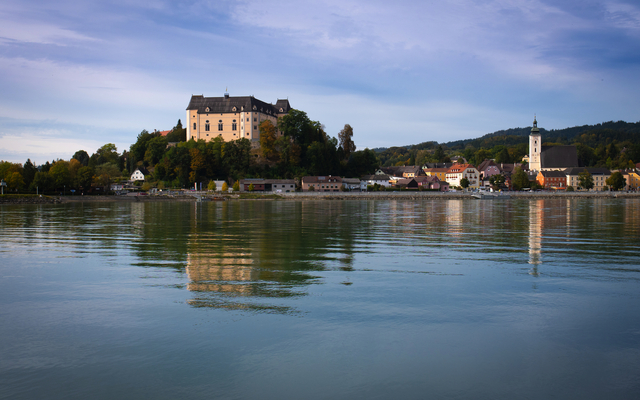 Flusslandschaft mit Schloss und Kirche im Hintergrund, sanfte Hügel und bewölkter Himmel.
